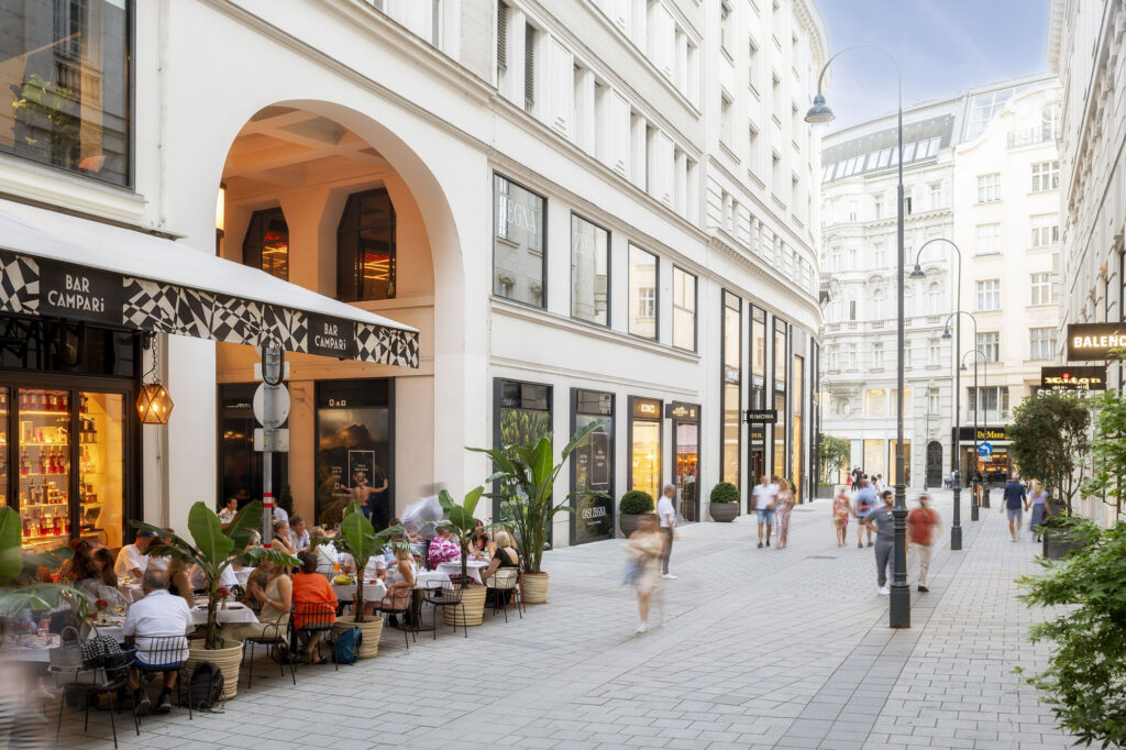 Pedestrian passage with café terrace under arches and shoppers walking past outdoor seating.